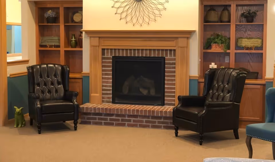 A cozy living room area with a brick fireplace centered between two wooden built-in shelves. Two black leather tufted armchairs are positioned on either side of the fireplace. Decorative items and plants are displayed on the shelves, and a sunburst wall decoration hangs above the fireplace.