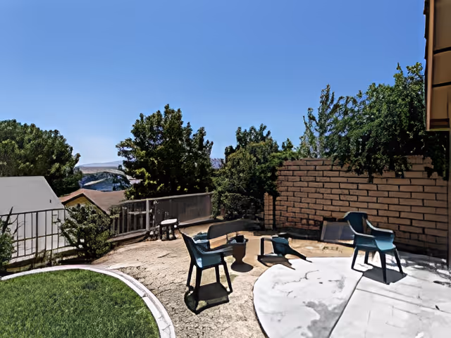 Backyard patio with plastic chairs around a small table and fire pit, a lawn area, brick wall and trees under a clear blue sky.