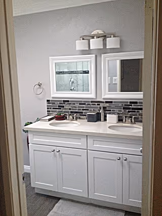 A bathroom with a double sink vanity featuring white cabinets and a white countertop. Above the sinks are two rectangular mirrors with white frames, and a modern light fixture with three lights is mounted on the wall. The backsplash consists of horizontal tiles in shades of gray, black, and white. A towel ring is mounted on the left wall, and a small plant and toiletries are placed on the countertop.