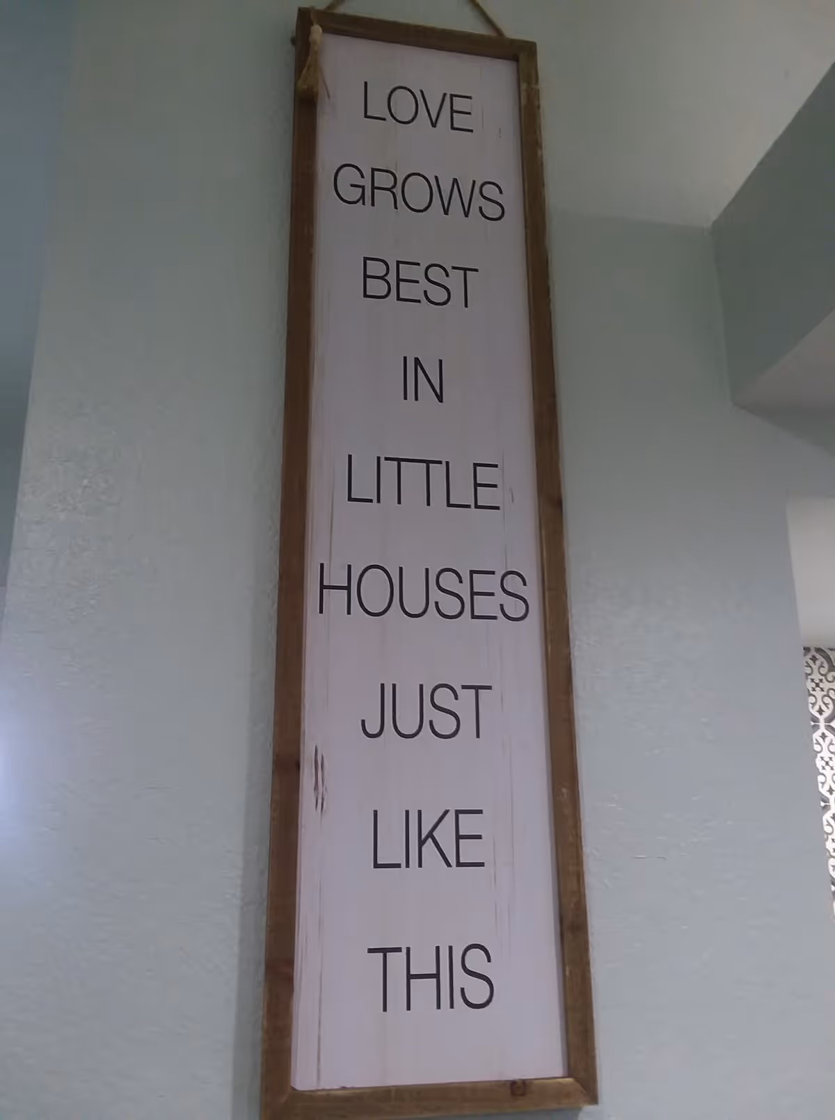Tall vertical wooden wall sign with the text "LOVE GROWS BEST IN LITTLE HOUSES JUST LIKE THIS" hung on an interior wall.