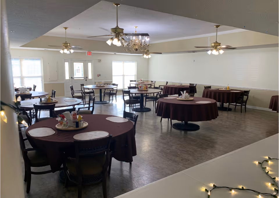 A dining room with multiple round tables covered with dark tablecloths and set with placemats and condiment trays. The room has ceiling fans with lights and a chandelier, large windows letting in natural light, and a door at the far end.