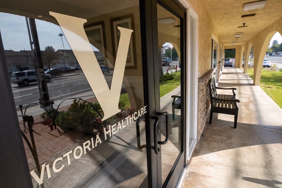 Glass entrance door marked "Victoria Healthcare" reflecting the street and leading to a covered walkway with benches.