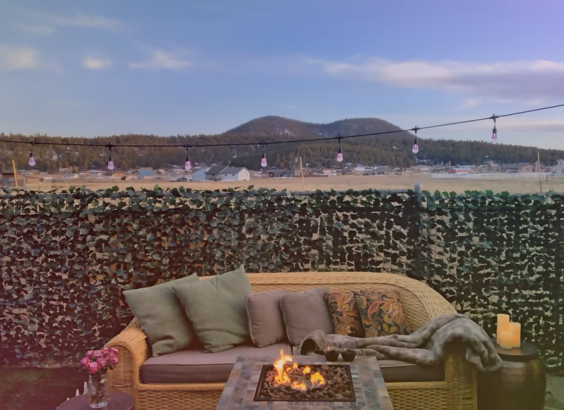 Outdoor patio area with a wicker sofa adorned with multiple cushions and a blanket. In front of the sofa is a fire pit table with flames burning. String lights hang above, and a privacy fence covered with greenery is in the background. Beyond the fence, there is a view of a rural landscape with houses, trees, and hills under a partly cloudy sky.