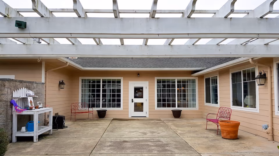 Outdoor patio area at McMinnville Memory Care with a concrete floor, beige walls, large windows, a white door in the center, two red metal benches, two large empty plant pots, and a white pergola overhead. There is a small white table with various items on the left side near a stone wall.