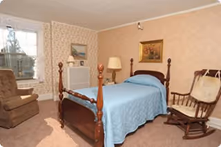 A cozy bedroom with a wooden bed frame covered with a light blue quilt. The room has beige wallpaper with a subtle pattern, a window with floral curtains, a brown armchair, a wooden rocking chair with a cushion, a floor lamp, and framed artwork on the walls.