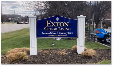Outdoor sign for Exton Senior Living, a personal care and memory care community, positioned on a grassy area near a road with trees and parked cars in the background.
