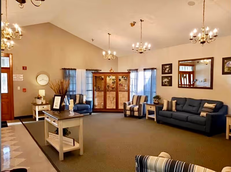 A spacious and well-lit living room area with beige walls and carpeted floor. The room features a blue sofa with striped pillows, two striped armchairs, a wooden display cabinet with glass doors, a large mirror on the wall, and several framed pictures. There are multiple chandeliers hanging from the ceiling, and a small table with decorative items near the entrance door.