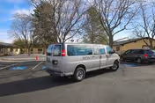 A white Monte Vista Hills transport van parked in a care facility parking lot with accessible spaces, nearby buildings and leafless trees.