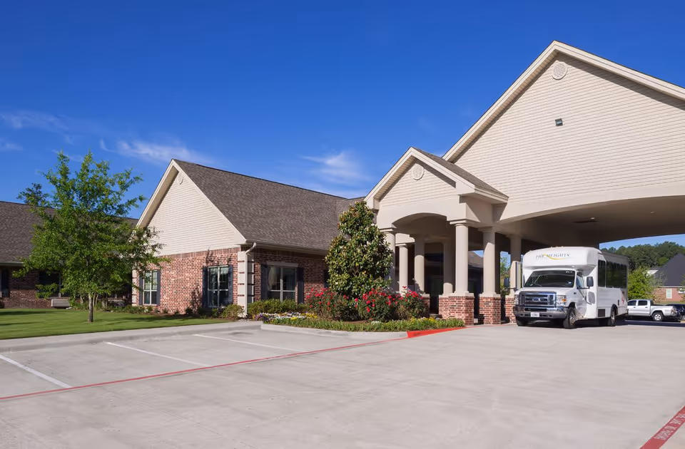 Front entrance of The Heights of Tyler senior living building with a covered porte-cochere, landscaped beds and a shuttle bus parked at the curb.