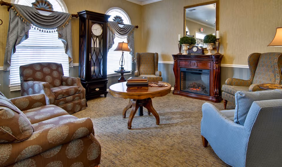 A cozy living room with patterned armchairs and a sofa arranged around a round wooden coffee table. The room features two arched windows with decorative curtains, a tall grandfather clock, a wooden electric fireplace with a mirror above it, and table lamps providing warm lighting.