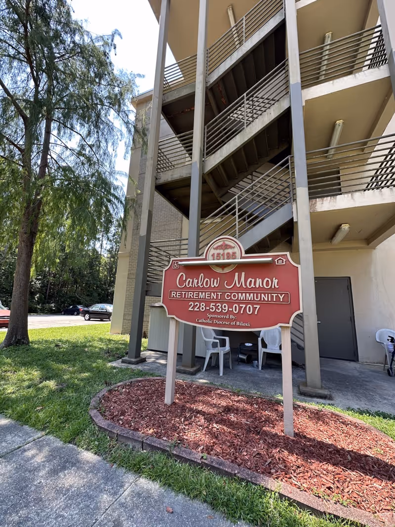 Front exterior of Carlow Manor Retirement Community showing the building entrance, metal stairwell, and a sign in a small landscaped area.
