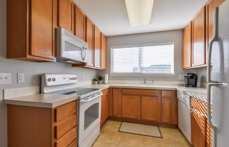 A bright kitchen with wooden cabinets, a white electric stove with oven, a microwave above the stove, a dishwasher, a refrigerator, a coffee maker, and a window with blinds letting in natural light.