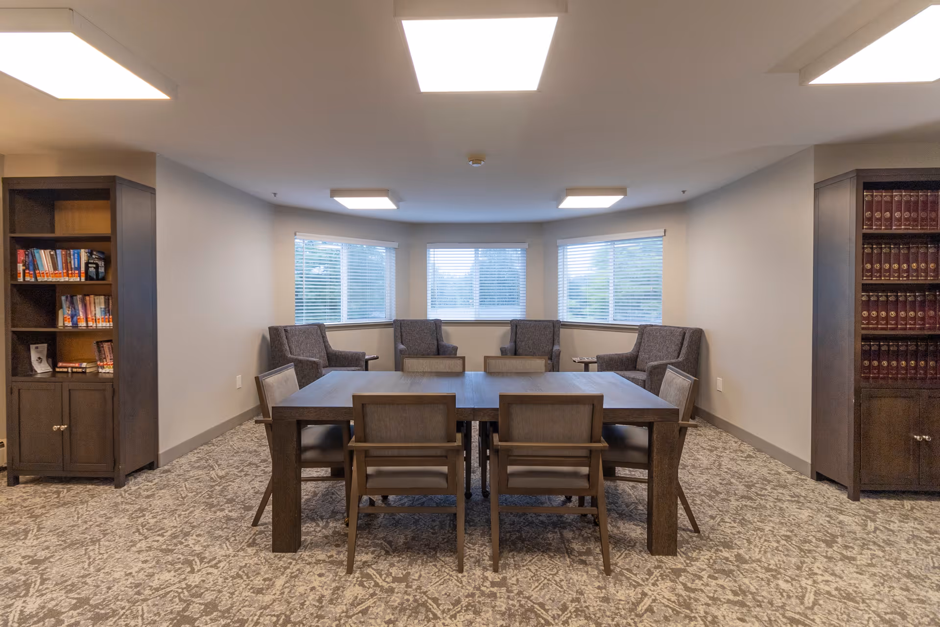 A quiet common area with a rectangular wooden table surrounded by six chairs in the center. Behind the table are four upholstered armchairs arranged near three large windows with blinds. On either side of the room are tall wooden bookshelves filled with books. The room has a patterned carpet and bright ceiling lights.