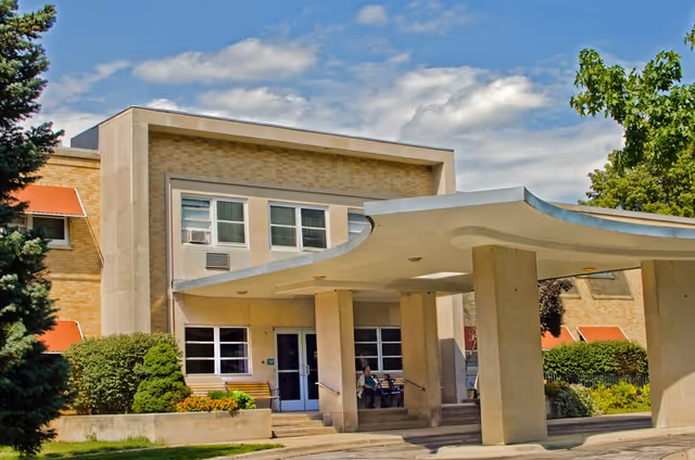 Exterior view of a senior living facility building with a covered entrance supported by large pillars. The building has beige brick walls, several windows, and red awnings. There are bushes and trees around the entrance area under a partly cloudy sky.
