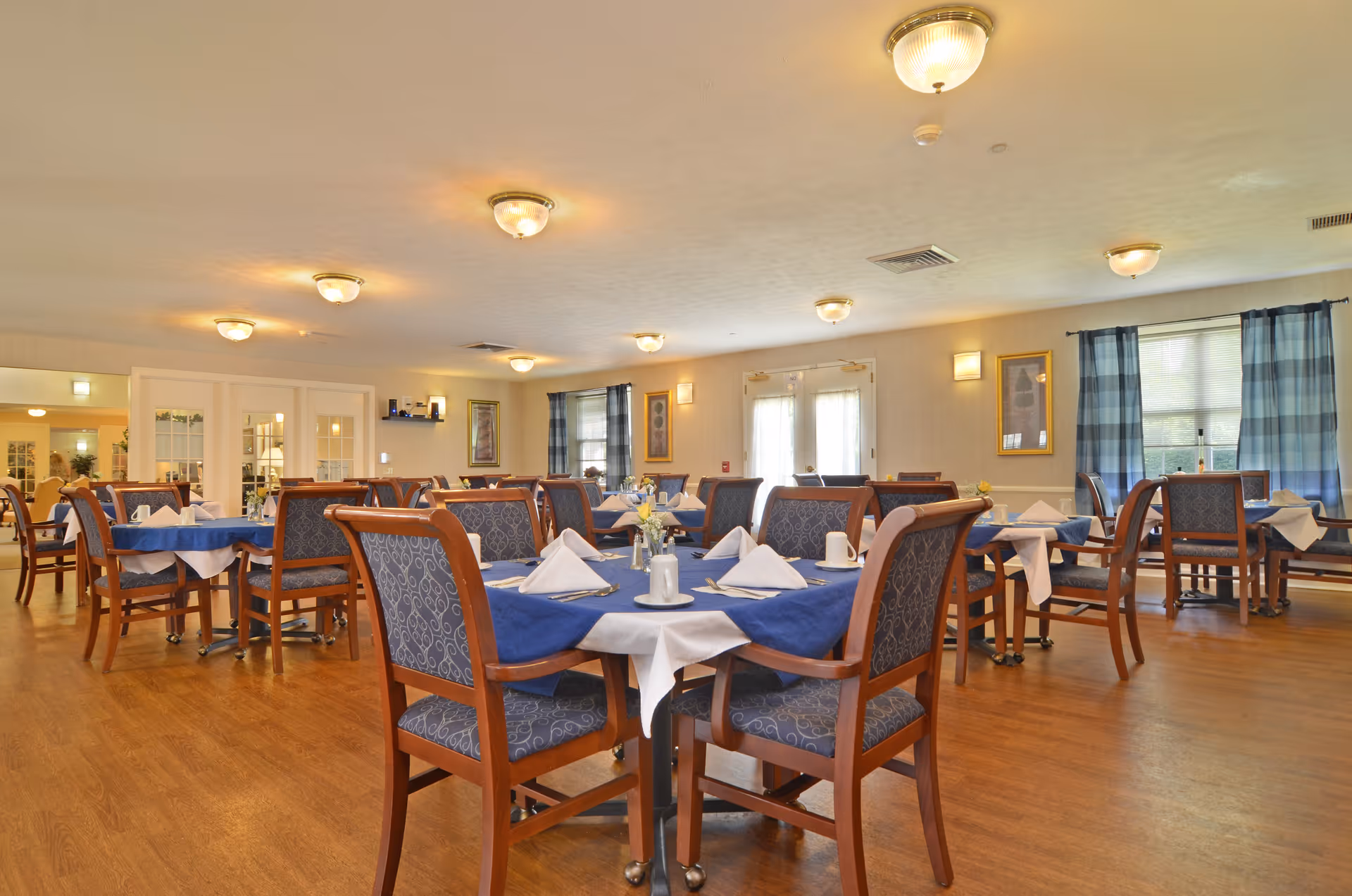 A spacious dining room with multiple wooden tables covered with blue and white tablecloths, each set with white napkins, cups, and small flower vases. The room has wooden flooring, several ceiling lights, windows with blue checkered curtains, and framed artwork on the walls.