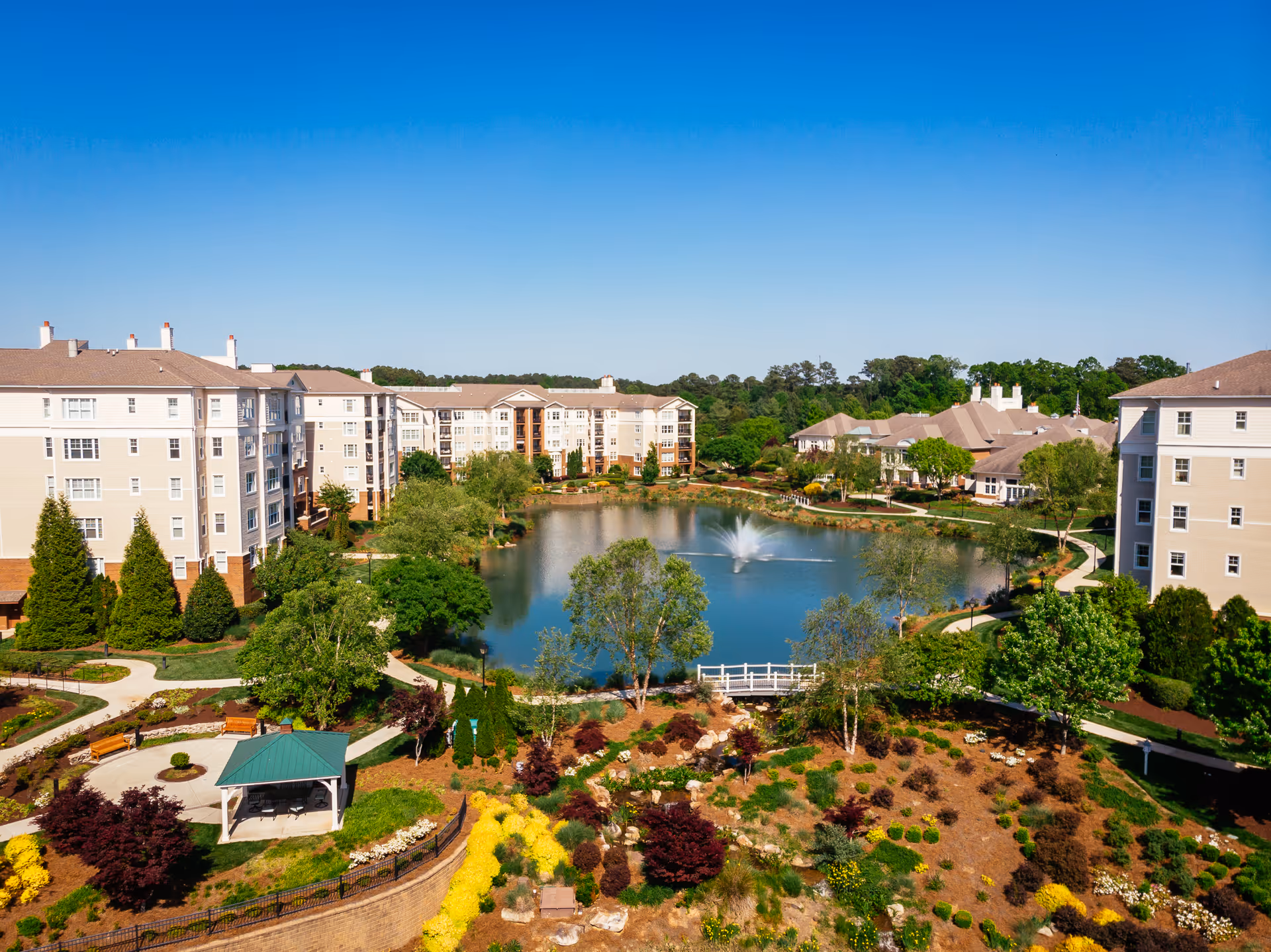 Aerial view of The Cypress Of Raleigh senior living facility showing multiple beige multi-story buildings surrounding a large pond with a fountain in the center. The landscaped grounds include walking paths, benches, a small gazebo with a green roof, and various trees and colorful shrubs under a clear blue sky.