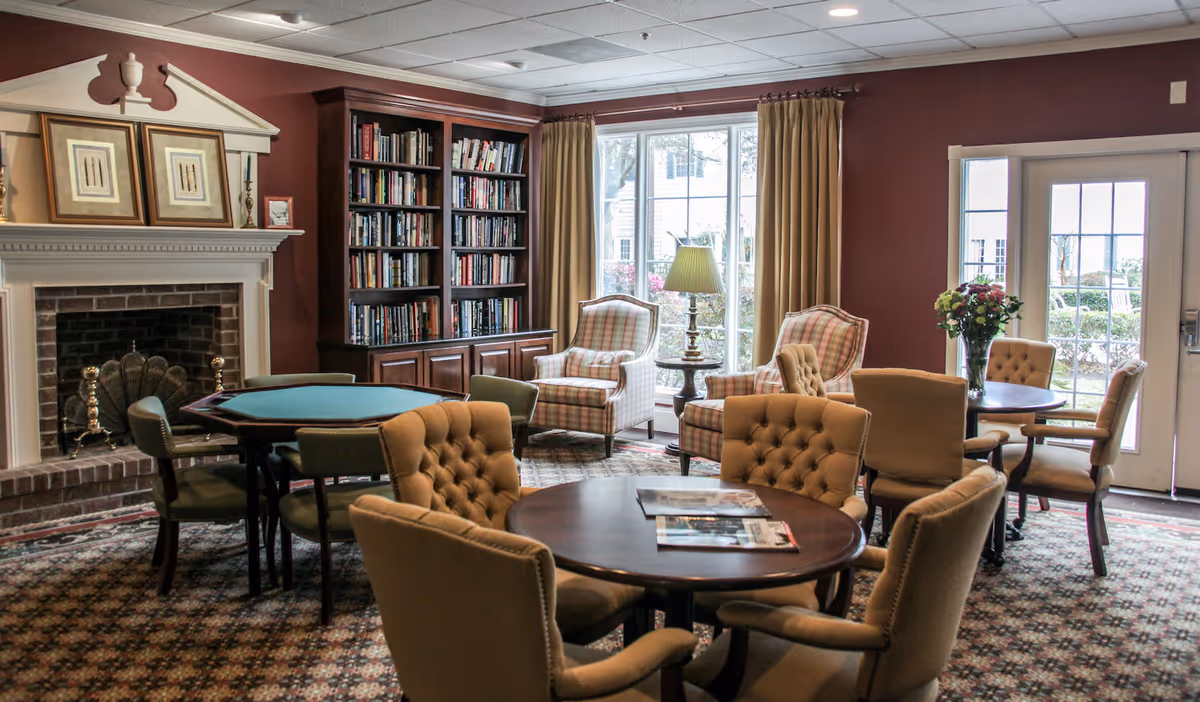 A cozy common area with multiple round tables surrounded by cushioned chairs. There is a fireplace with framed artwork above it on the left side, a bookshelf filled with books, and two plaid armchairs near large windows with beige curtains. A lamp and a vase with flowers are also visible, with glass doors leading outside.