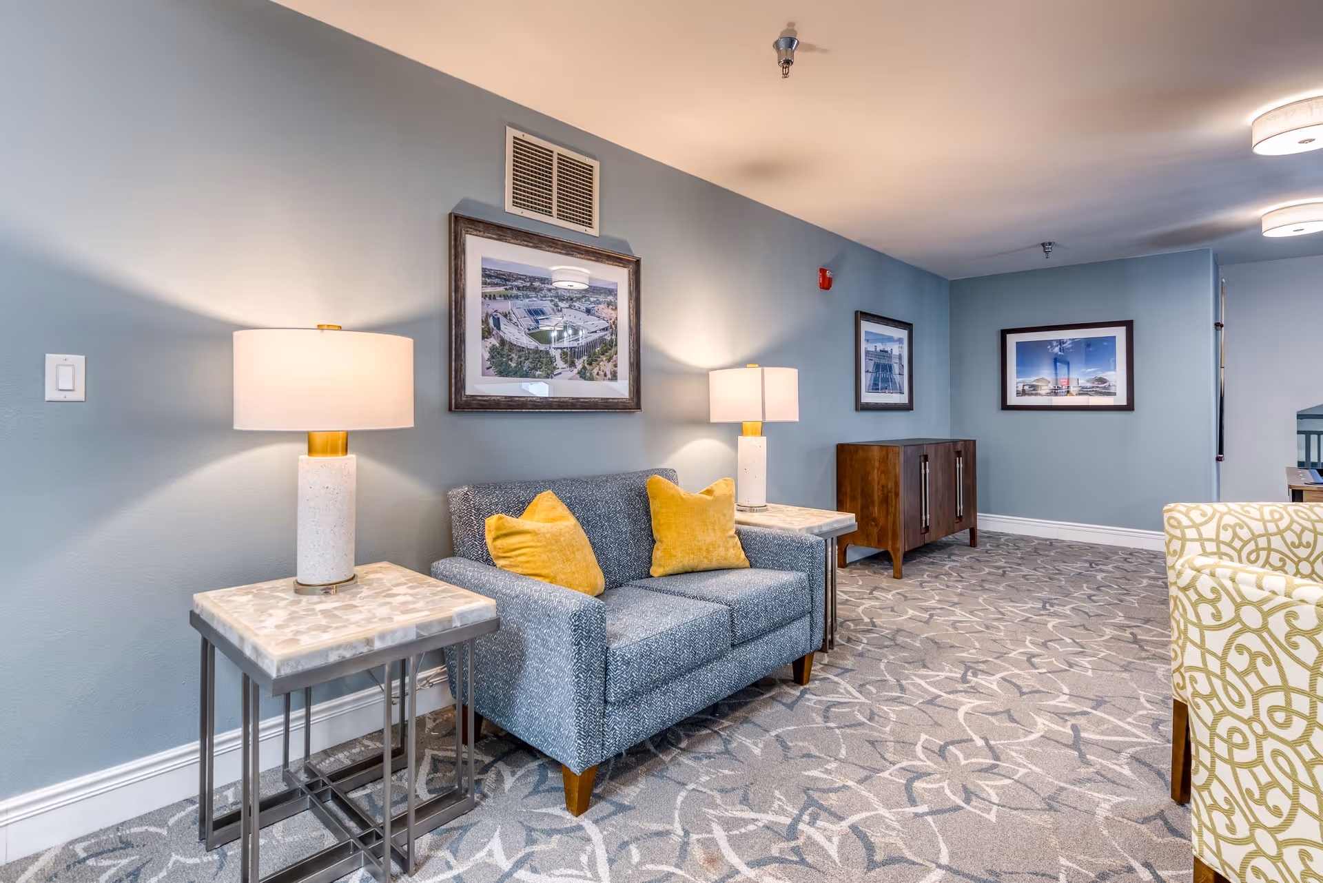 A cozy seating area in a senior living facility with a gray upholstered loveseat adorned with two yellow pillows, flanked by two side tables each holding a lamp with a white shade. The walls are painted light blue and decorated with framed pictures. The floor is carpeted with a floral pattern, and there is a wooden cabinet against the wall in the background.