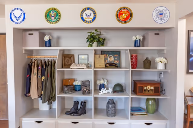 A white shelving unit displaying military memorabilia including jackets, boots, framed certificates, a helmet, decorative vases, and plants. Above the shelves are five circular emblems representing different branches of the United States military: Air Force, Army, Navy, Marine Corps, and Coast Guard.