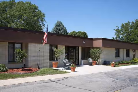Front exterior of a single-story senior living facility with flag and potted plants.