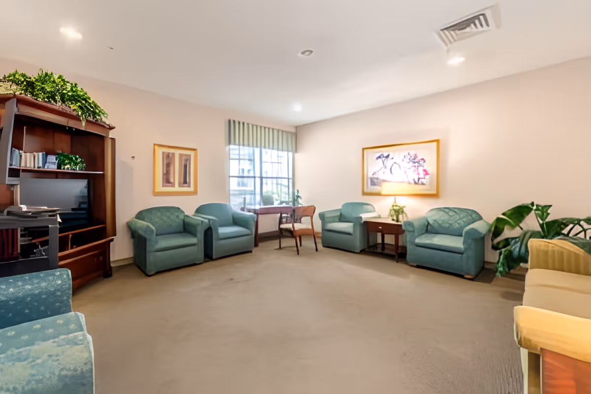 Bright lounge with green upholstered armchairs arranged around a central carpeted area, a TV cabinet, side tables, plants and framed artwork.