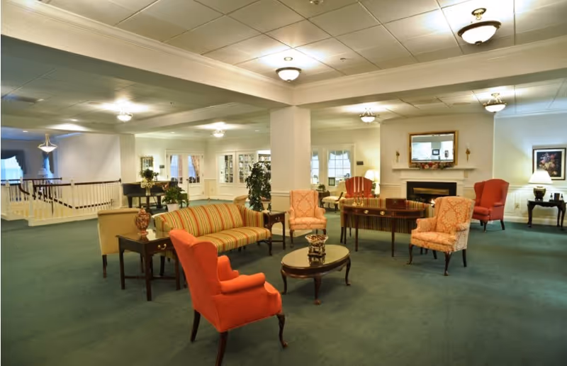 Spacious lounge in a senior living facility with upholstered sofas and armchairs arranged around coffee tables and a fireplace.