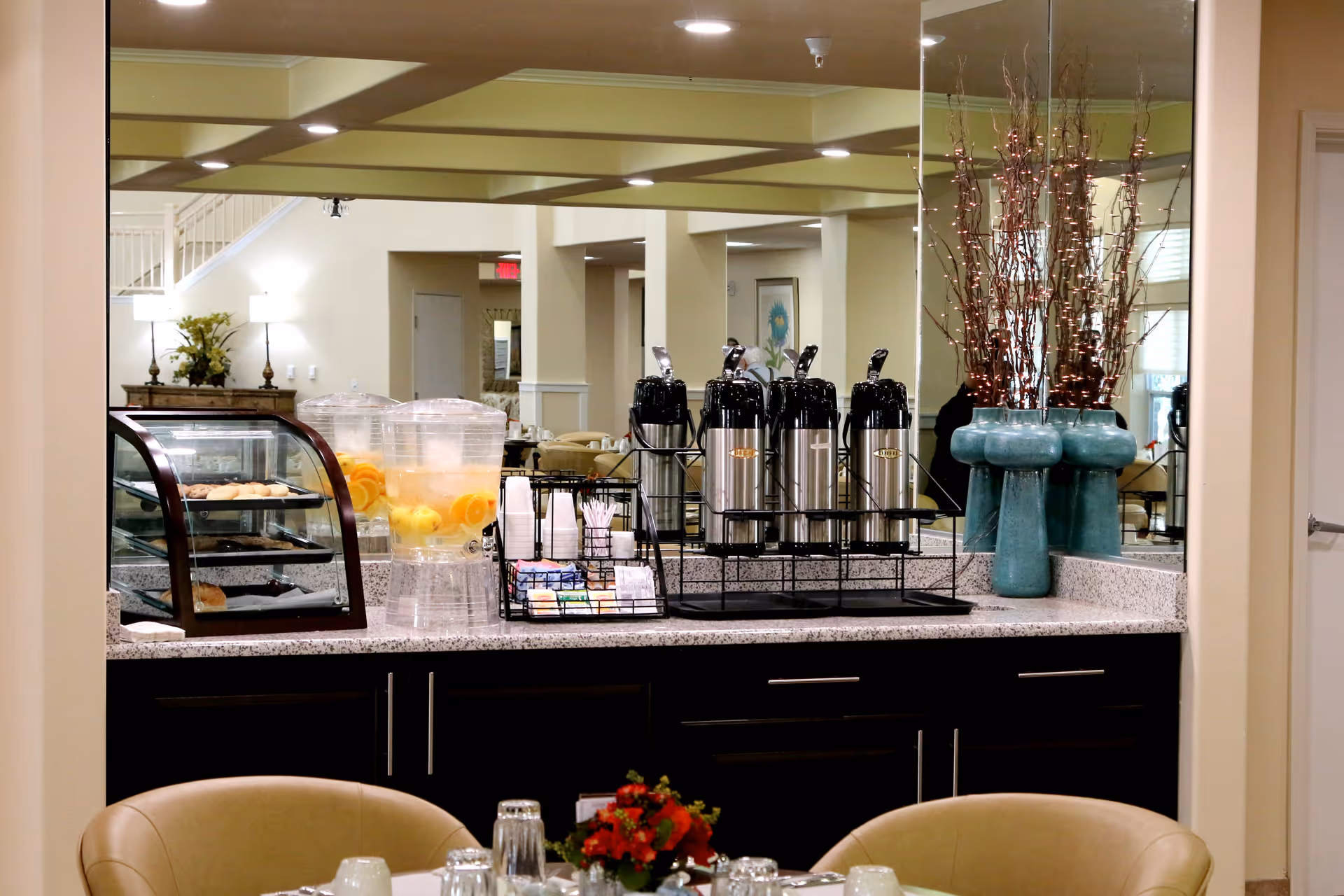 A beverage and snack station in a dining area featuring a countertop with coffee dispensers, a water dispenser with lemon and orange slices, a display case with pastries, and a rack with cups and condiments. In the foreground, a table is set with glasses, cups, and a small floral centerpiece. The background shows a well-lit room with light-colored walls, a staircase, and decorative vases with branches.