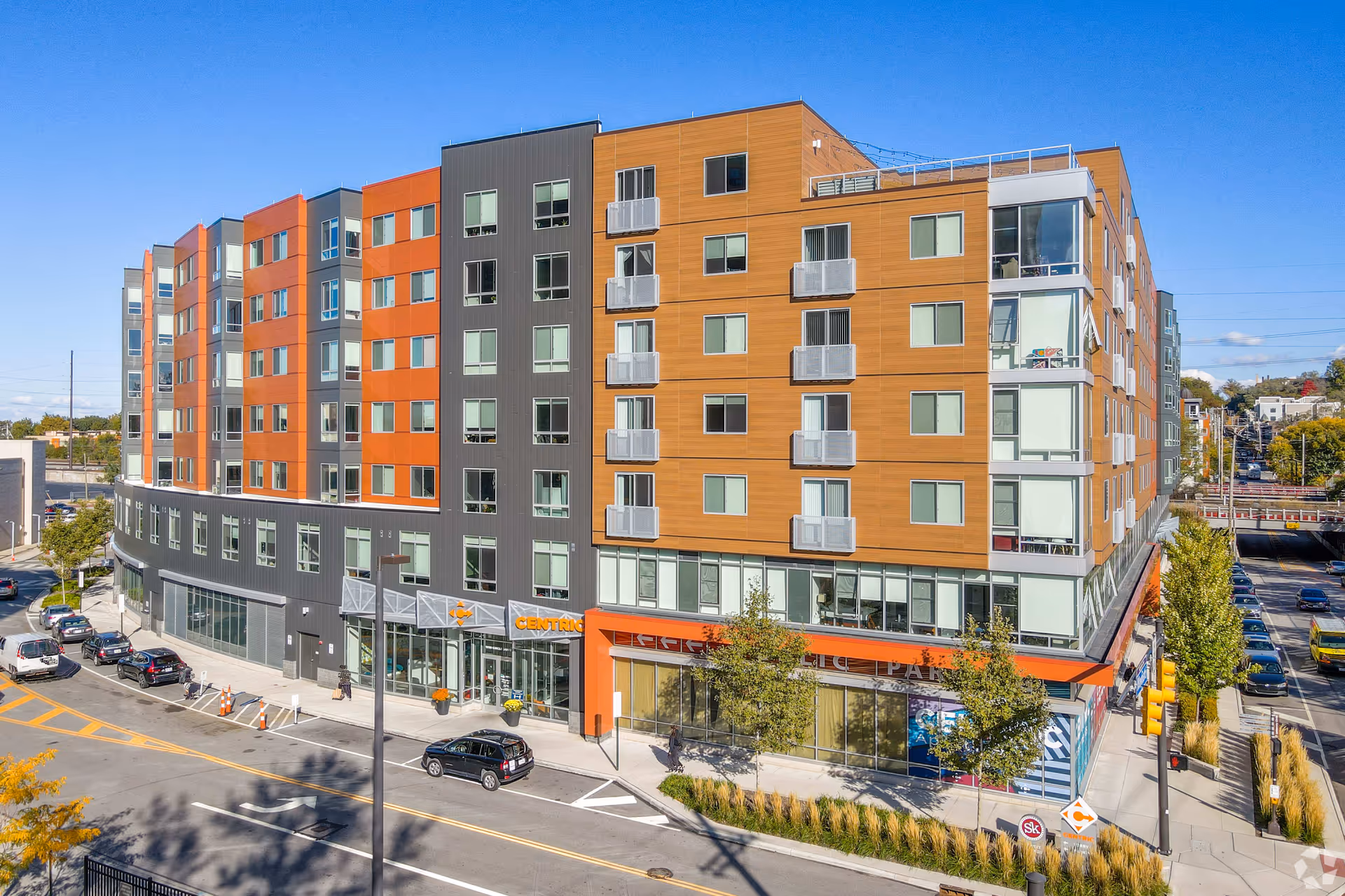 Exterior view of a modern multi-story residential building with orange, gray, and wood-toned panels. The building has many windows and small balconies, with a street and parked cars in front. Trees and a clear blue sky are visible.