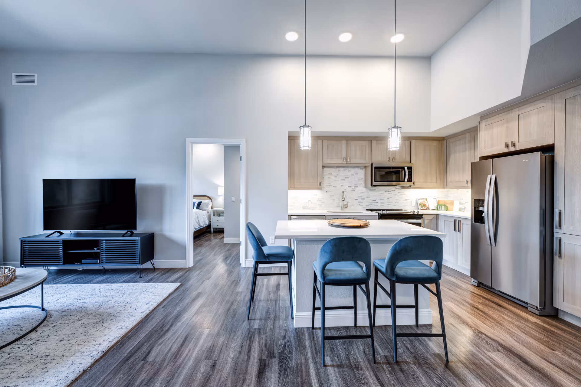 Modern open-concept living space featuring a kitchen with light wood cabinets, stainless steel refrigerator, microwave, and stove. A white island with three blue upholstered bar stools is in the center. To the left, there is a living area with a flat-screen TV on a black media console and a light-colored rug on wood flooring. A doorway leads to a bedroom with a bed visible in the background.