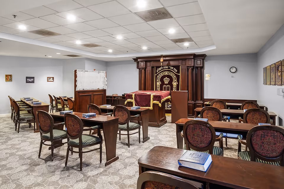 Interior view of a synagogue or prayer room with wooden desks and chairs arranged in rows. The room features a large wooden ark with a decorative curtain and Hebrew inscriptions at the front. The walls are light-colored with framed pictures, and the ceiling has recessed lighting.