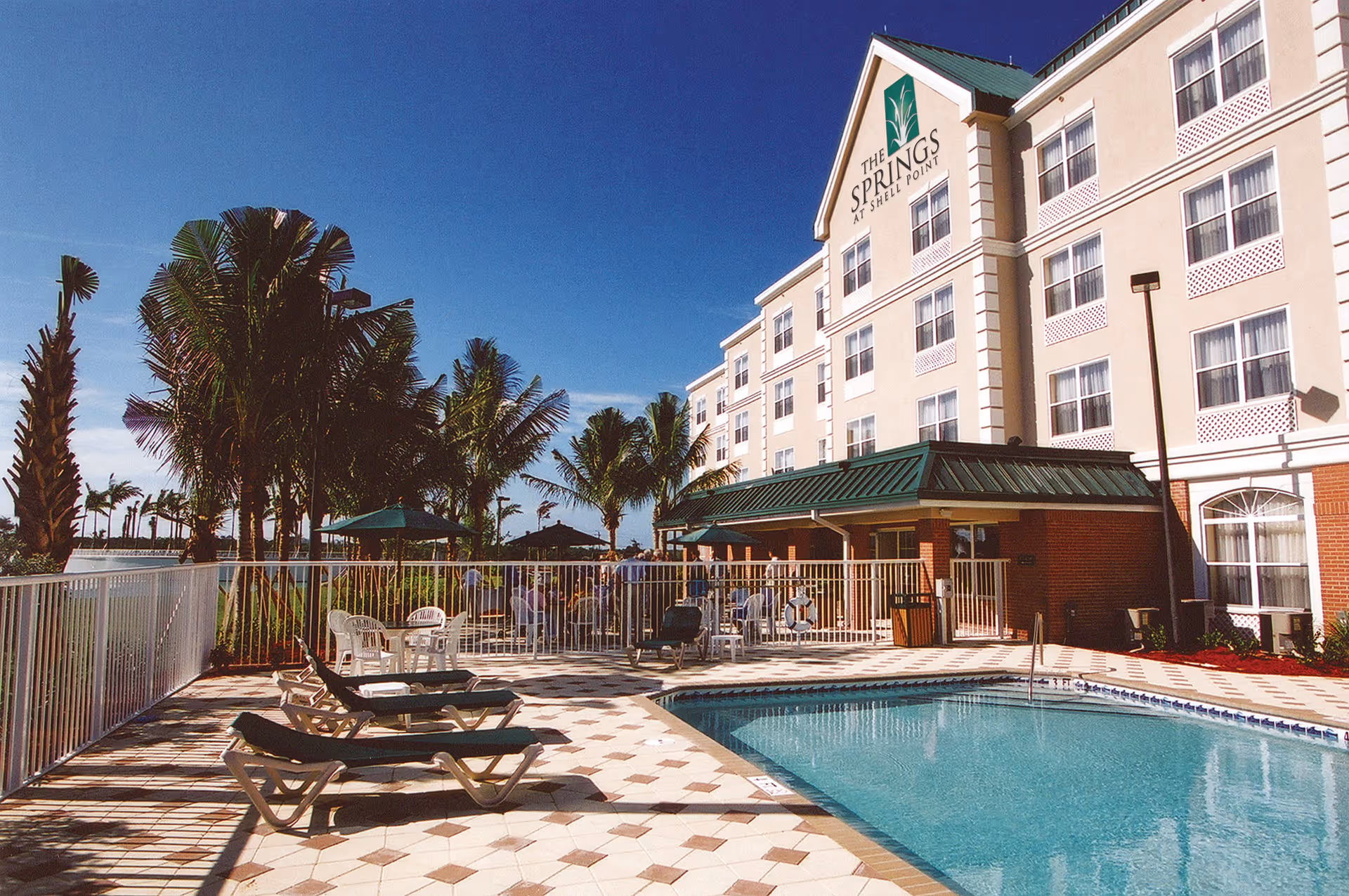 Outdoor swimming pool and sun loungers beside a multi-story senior living building with palm trees under a clear blue sky.