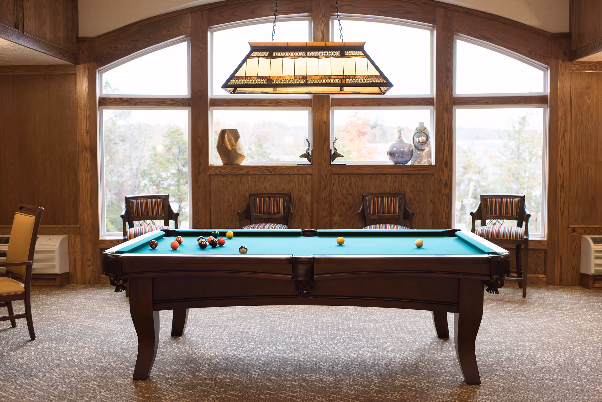 A pool table with billiard balls arranged on the green felt surface in a room with large windows and wooden paneling. Four wooden chairs with striped cushions are placed against the wall beneath the windows, and decorative vases and sculptures are displayed on the window ledge. A rectangular hanging light fixture is suspended above the pool table.