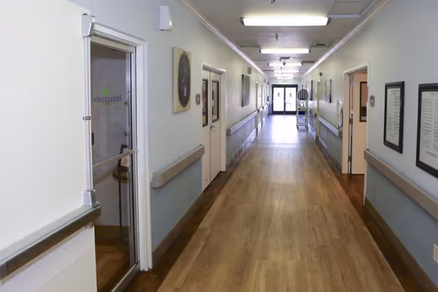 A long, clean hallway inside a healthcare facility with light-colored walls, wooden flooring, handrails on both sides, several doors along the corridor, and a glass exit door at the far end letting in natural light.