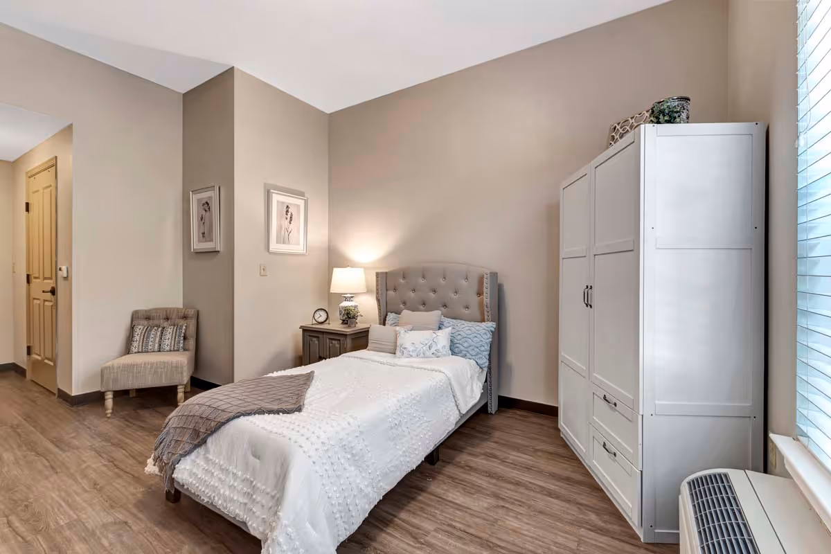 A neatly arranged bedroom with a single bed featuring a tufted headboard, white bedding, and multiple pillows. Next to the bed is a nightstand with a lamp and a small clock. A beige upholstered chair with a decorative pillow is placed near the doorway. A white wardrobe stands against the wall near a window with blinds, and the room has wood flooring and beige walls.