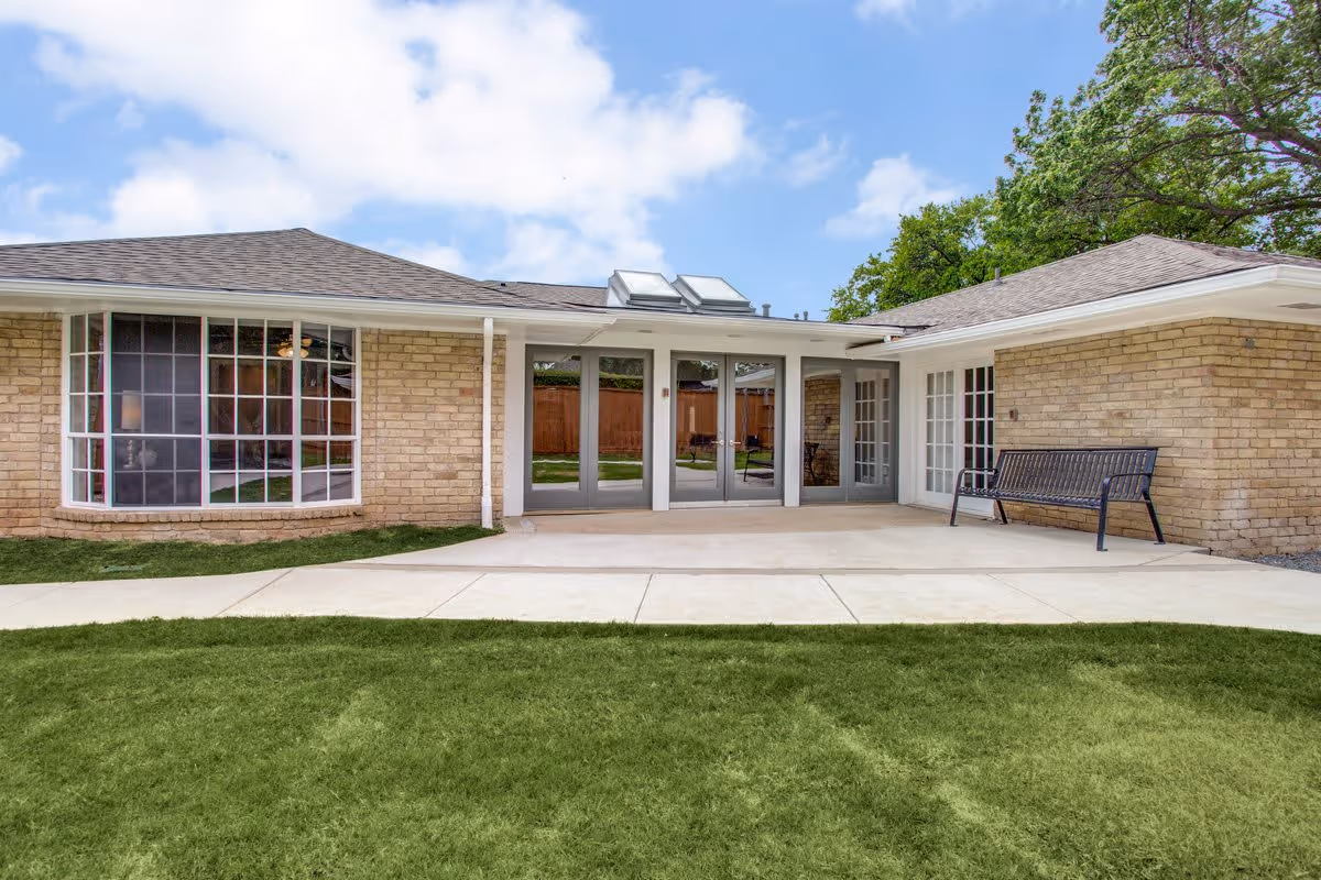 Exterior view of a single-story brick building with large windows and glass doors leading to a concrete patio area. There is a black metal bench on the right side of the patio, green grass in the foreground, and trees in the background under a partly cloudy sky.