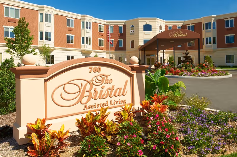 Front entrance of The Bristal Assisted Living with its stone sign, colorful landscaping, and covered porte-cochère.