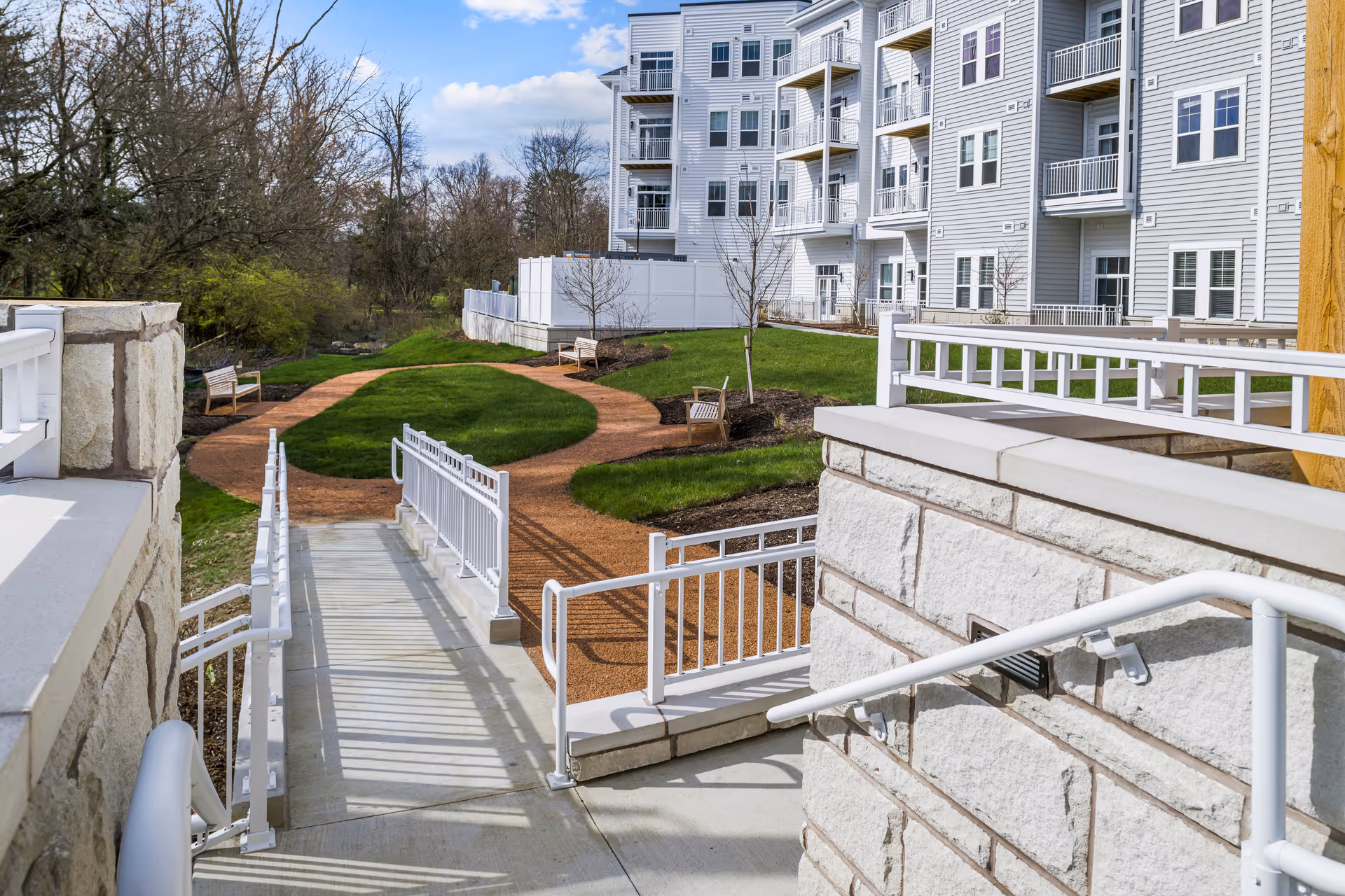 Outdoor view of a senior living facility with a paved walkway, benches, green grass, and a multi-story white building with balconies in the background under a partly cloudy sky.