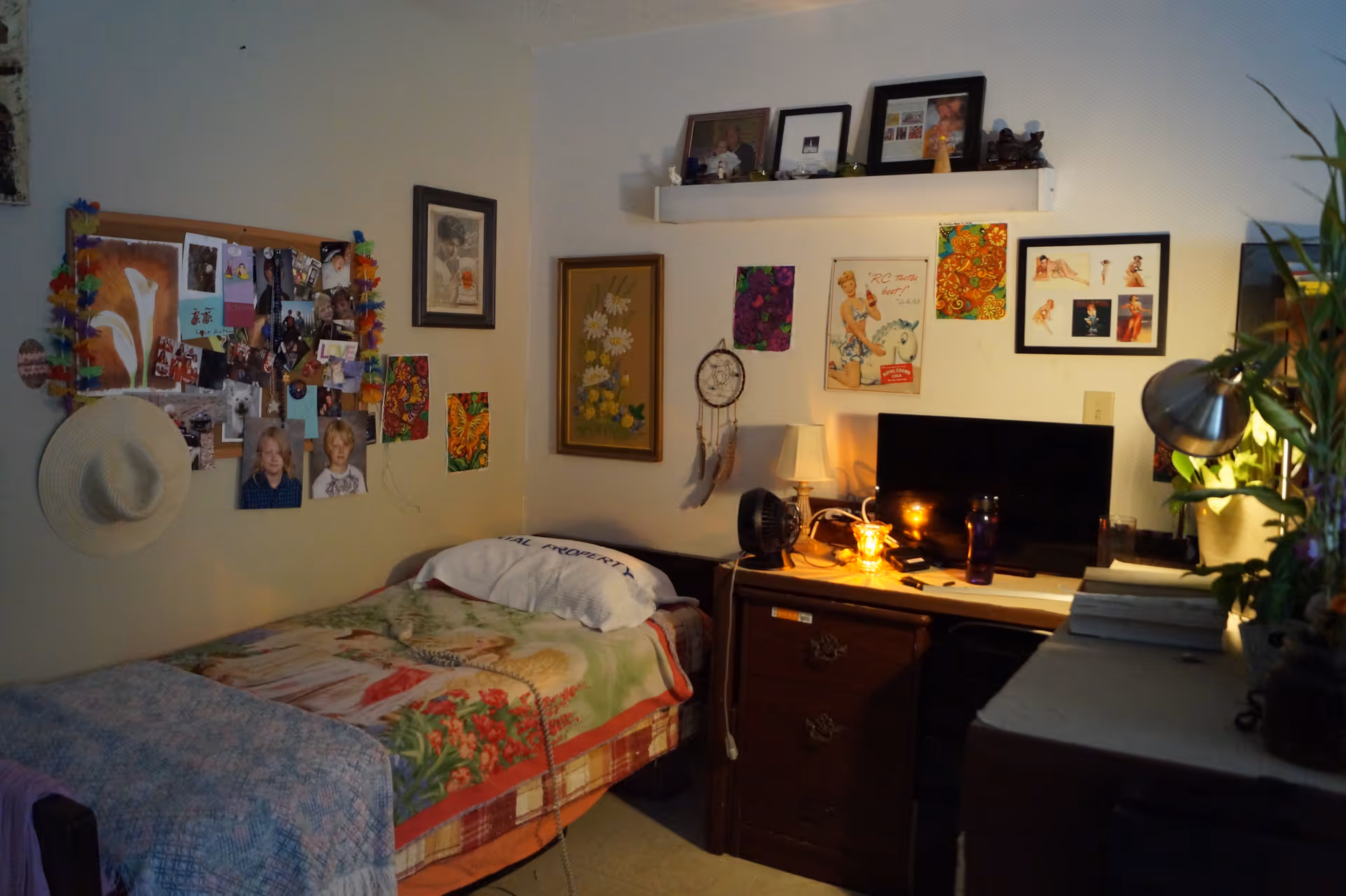 A cozy bedroom with a single bed covered in a colorful blanket and a pillow. The walls are decorated with various pictures, artwork, and a dreamcatcher. A desk with a lamp, a small fan, a computer monitor, and other personal items is next to the bed. A hat hangs on the wall near a bulletin board filled with photos and notes. There is a plant and a desk lamp on the right side of the image.