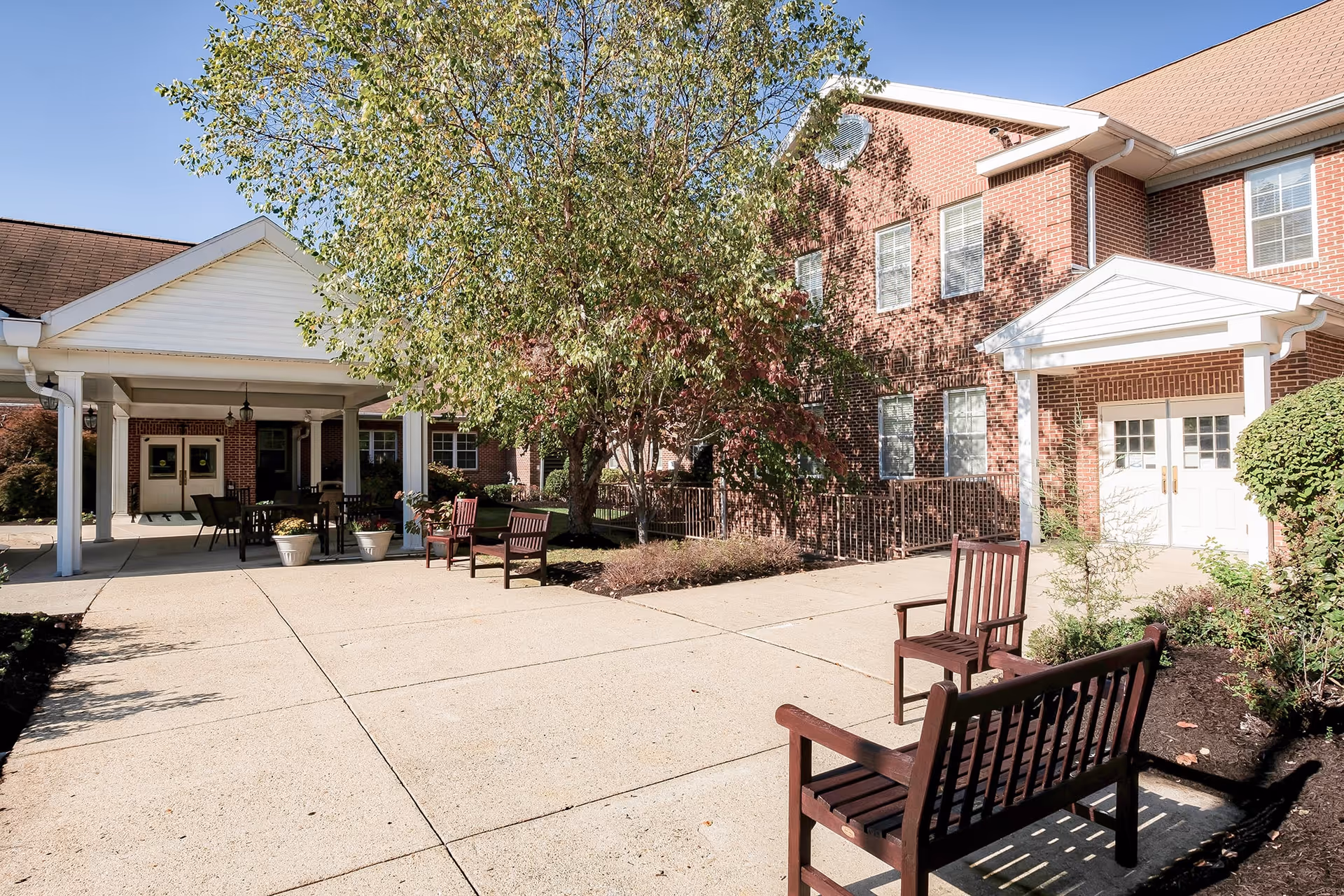 Outdoor courtyard area of a senior living facility with wooden benches, a large tree, potted plants, and a covered seating area. The building is made of red brick with white trim and multiple windows under a clear blue sky.