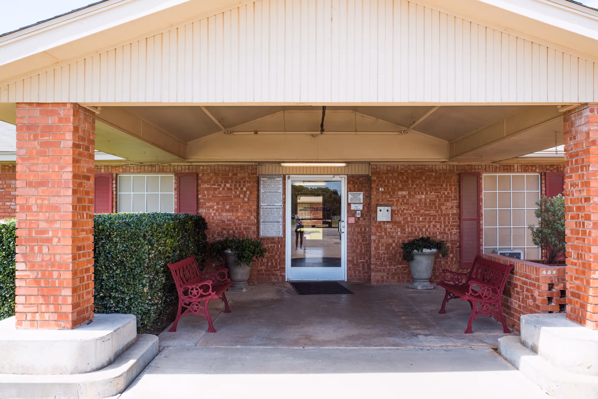 Entrance to a brick building with a covered porch supported by brick columns. There are two red benches on either side of the entrance, some potted plants, and windows with red shutters. The door is glass with a reflection of the outside visible.