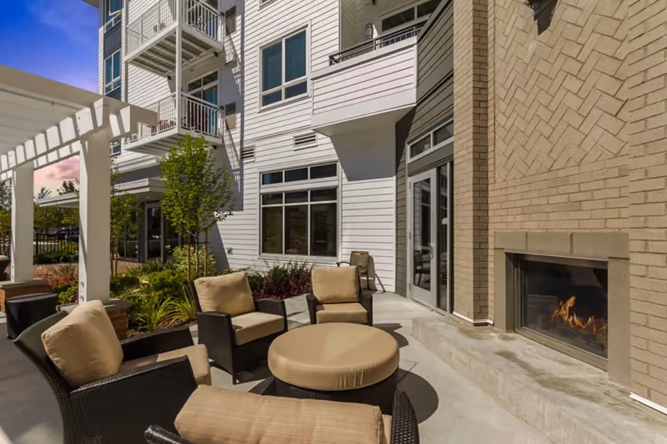 Outdoor patio area at Merrill Gardens at Burien featuring cushioned wicker chairs and a round ottoman arranged around a built-in outdoor fireplace, with a white pergola and landscaping including small trees and plants, adjacent to a multi-story building with balconies and large windows.