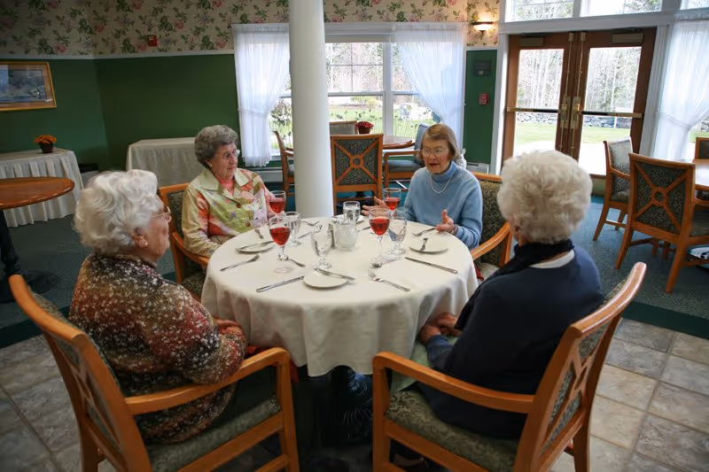Four elderly women sitting around a round dining table covered with a white tablecloth in a well-lit room with large windows and floral wallpaper. The table is set with glasses of red beverage, water glasses, plates, and silverware. The women appear to be engaged in conversation.