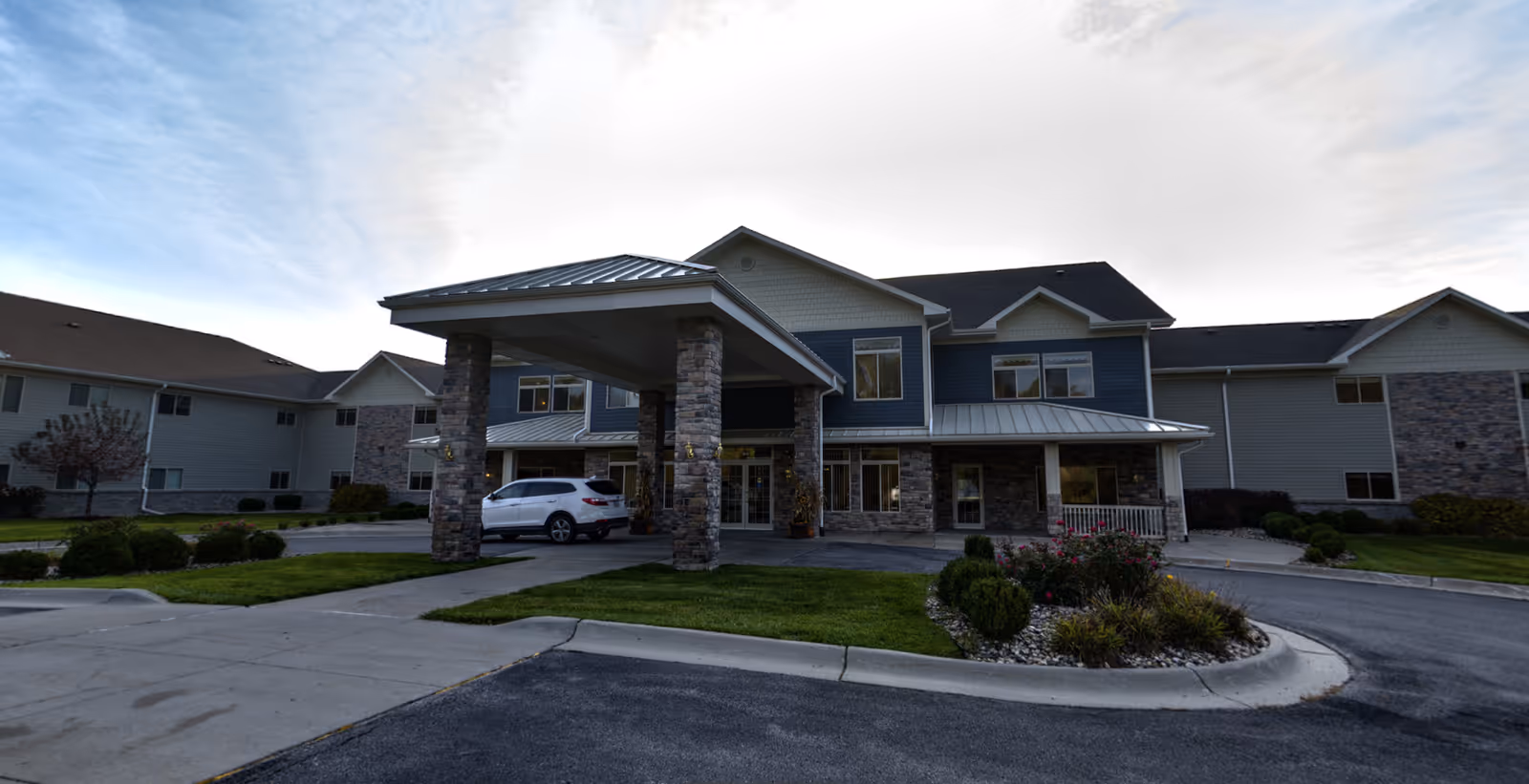 Front entrance of a two-story retirement community building with a covered porte-cochère, stone columns, landscaped circular driveway, and a parked white car.