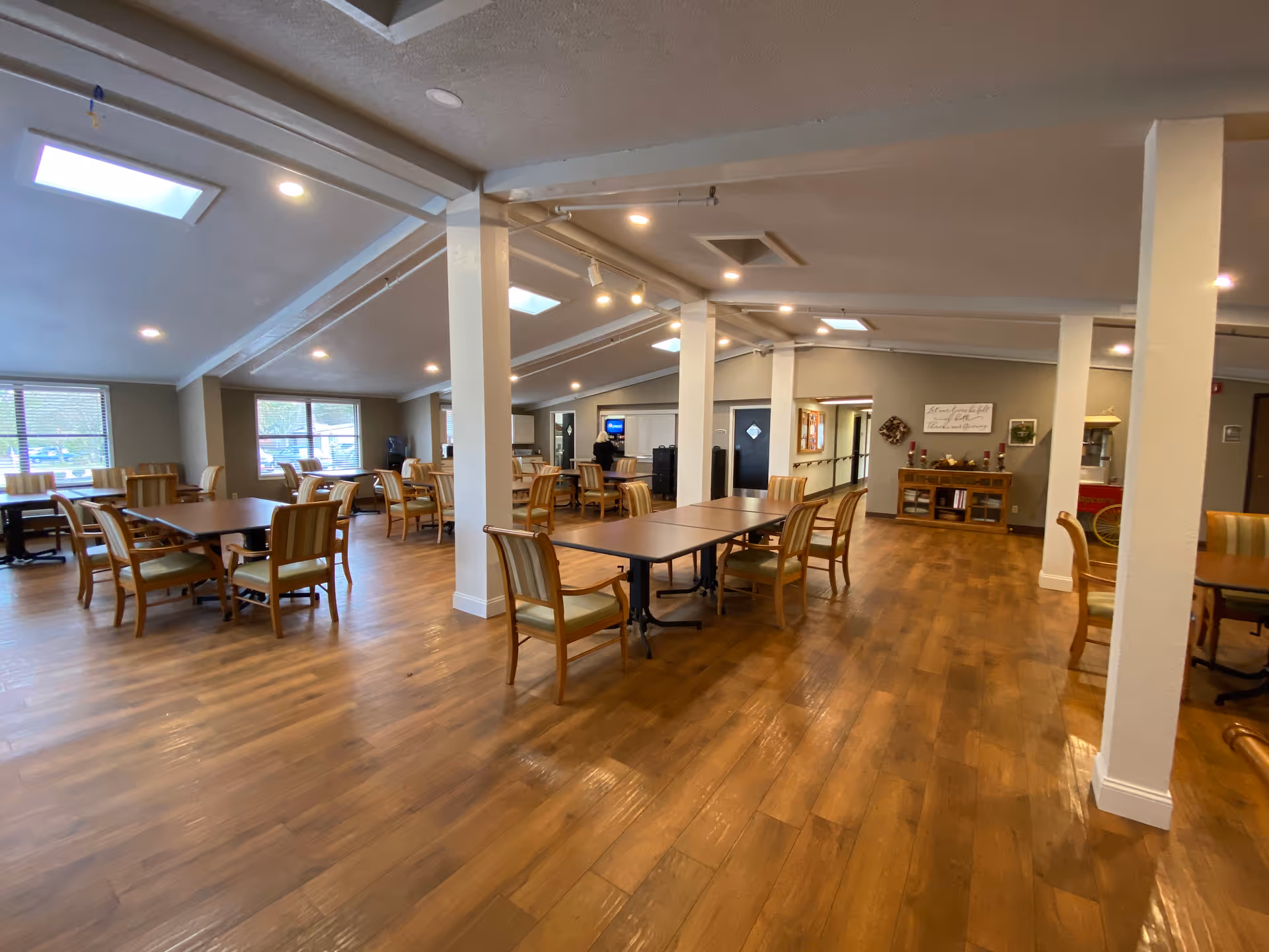 Spacious dining room with multiple wooden tables and chairs arranged neatly on a wooden floor. The room has large windows letting in natural light, recessed ceiling lights, and skylights. There are white support columns and a sideboard with decorative items and a sign on the wall in the background.