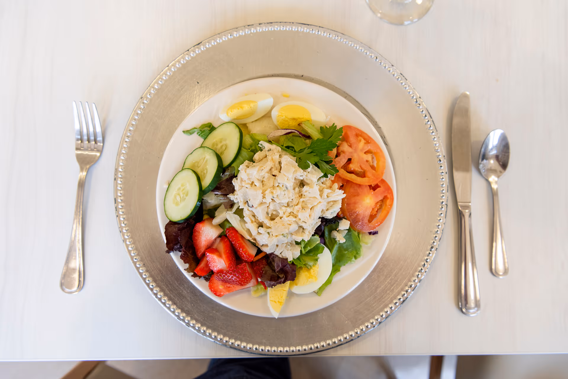 Top-down view of a plated salad with cucumbers, tomatoes, strawberries and hard-boiled eggs on a table with utensils.