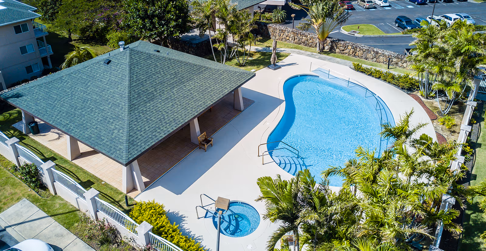 Aerial view of an outdoor pool area at The Ivy at Hawaii Kai featuring a kidney-shaped swimming pool, a smaller circular hot tub, a covered pavilion with a green roof, surrounded by palm trees and landscaping, with a parking lot visible in the background.