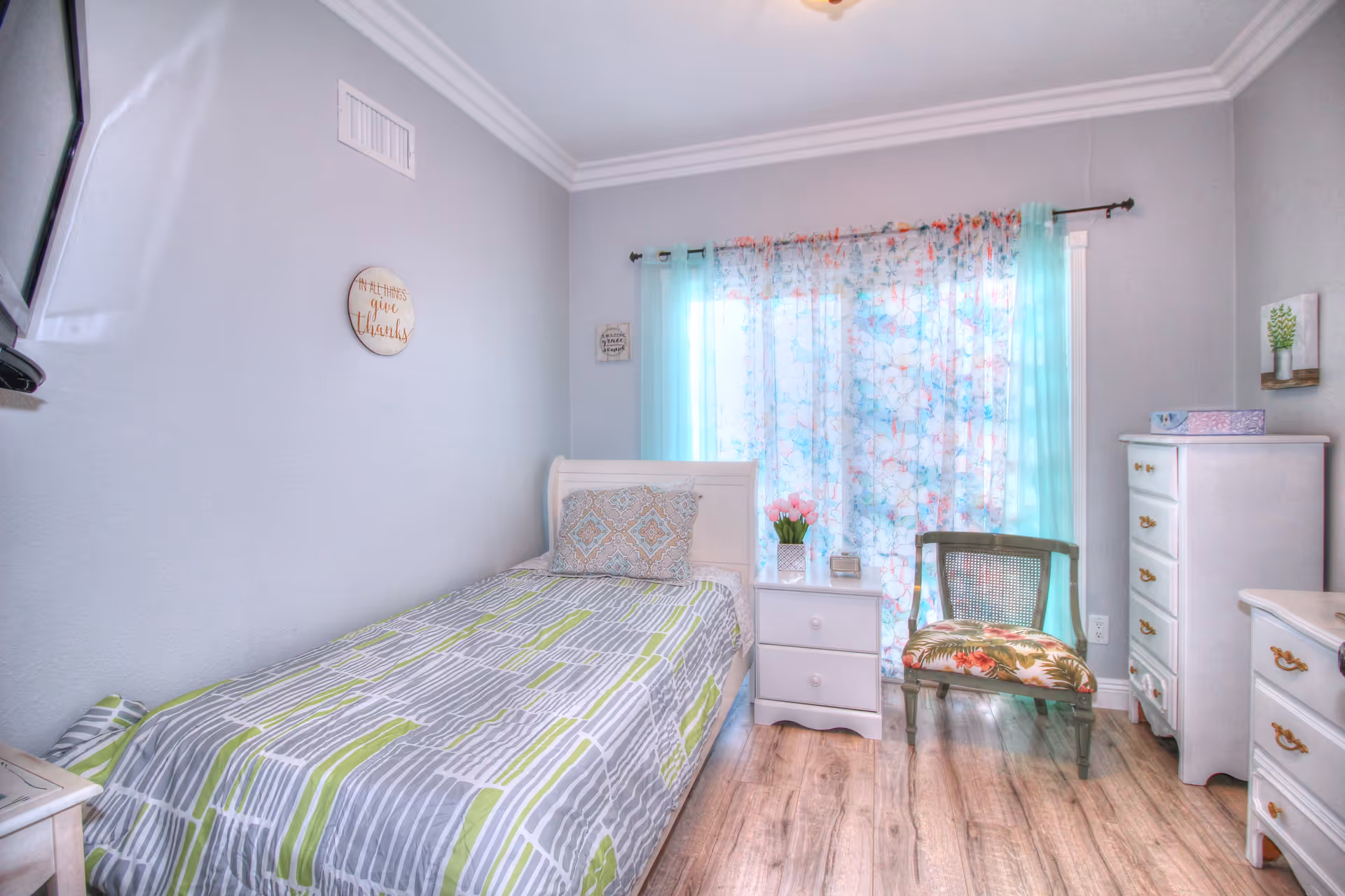 A small, cozy bedroom with a single bed covered in a gray, white, and green patterned bedspread. There is a decorative pillow on the bed. Next to the bed is a white nightstand with two drawers, a vase of pink flowers, and a small clock. A floral curtain with blue and pink hues covers the window behind the nightstand. To the right of the nightstand is a wooden chair with a floral cushion. The room also contains a white dresser and a chest of drawers with gold handles. The walls are painted light gray, and the floor is wooden. A round wall decoration with the words 'It is better to give thanks' is hung above the bed.
