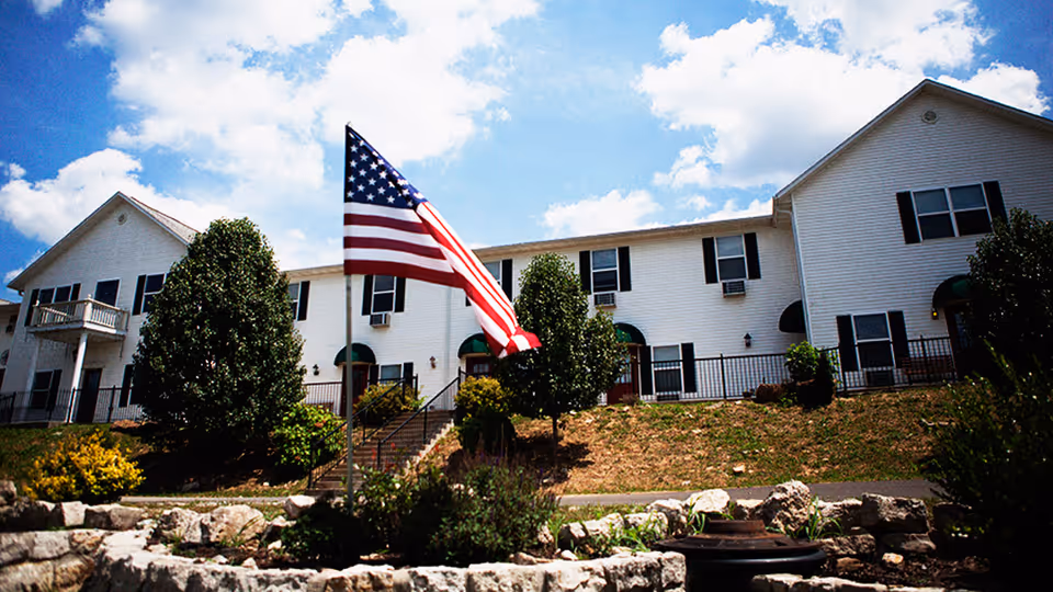 Two-story white assisted living building with an American flag and landscaped front lawn under a partly cloudy sky.