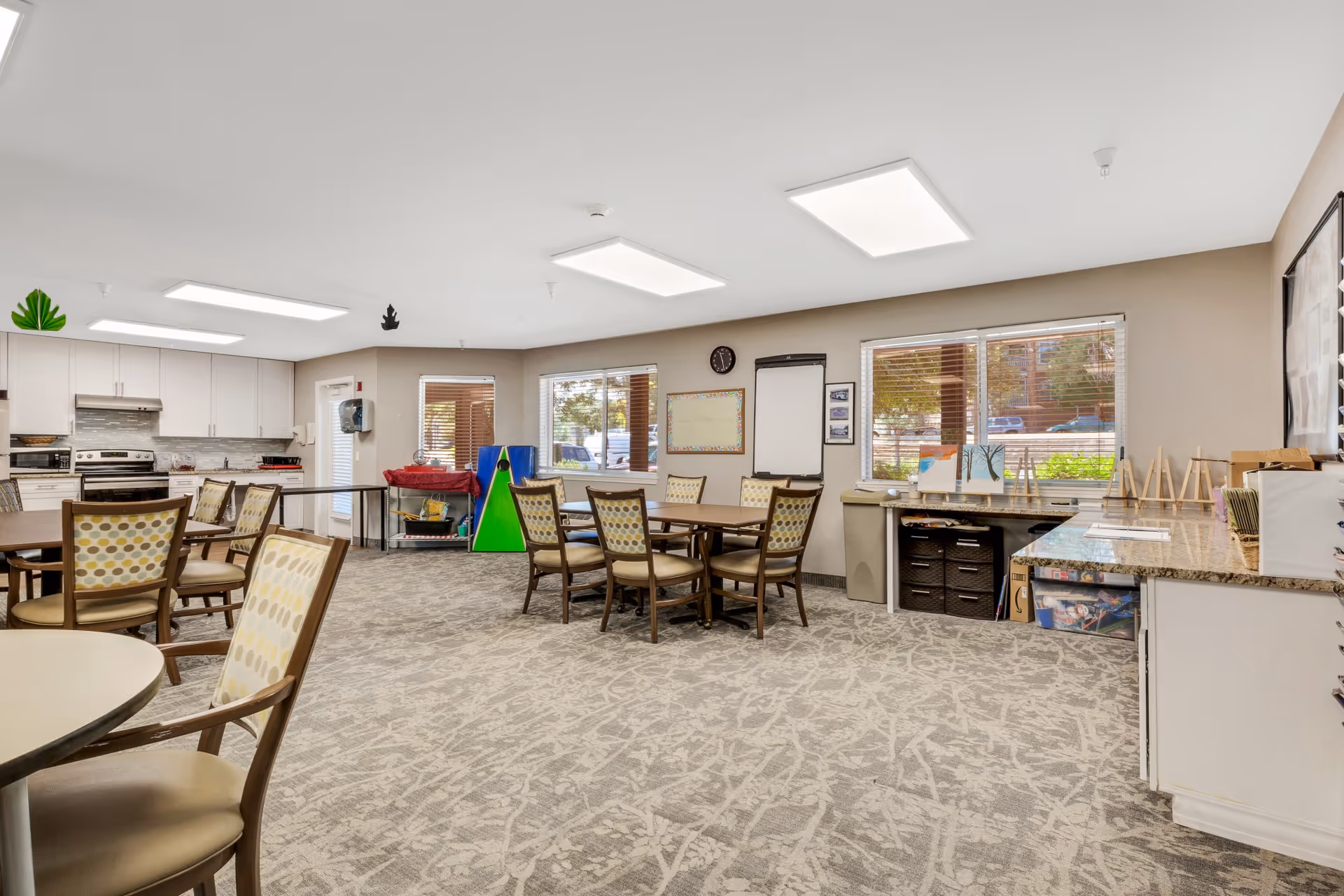 A spacious, well-lit activity room with several tables and chairs arranged for group activities. The room features a kitchenette area with a stove, microwave, and cabinets on the left side. There are windows with blinds letting in natural light, a clock on the wall, and various art supplies and easels on the right side near a countertop. The carpet has a light patterned design, and the ceiling has recessed lighting panels.
