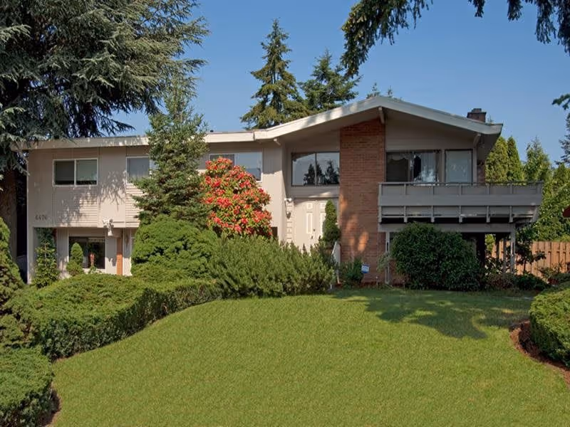A two-story residential building with beige siding and a brick chimney, surrounded by green shrubs, trees, and a well-maintained lawn under a clear blue sky.